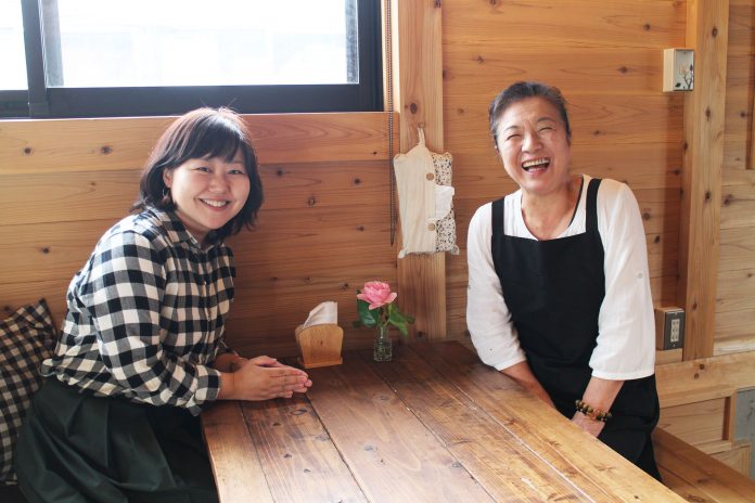 木の壁とテーブルに囲まれた温かみのある店内で、リポーターの女性と白いシャツに黒いエプロン姿の成澤さんが向かい合って笑顔で座っている写真