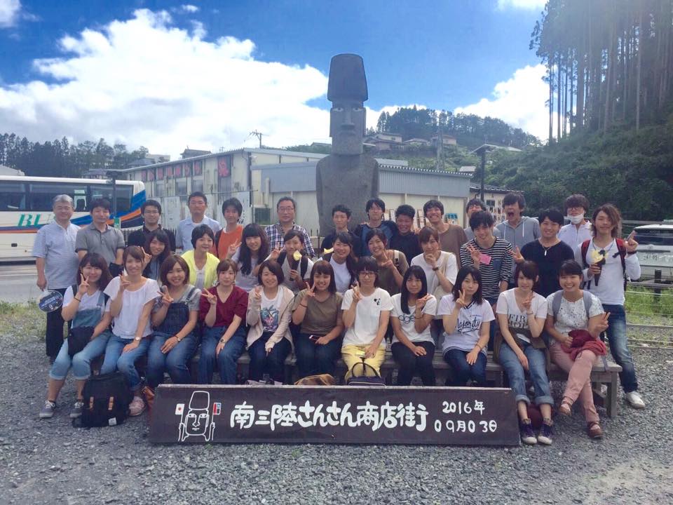 「南三陸さんさん商店街」と書かれた看板の後ろに商店街とモアイ像を背景にたくさんの若者たちが3列に並んでいる集合写真