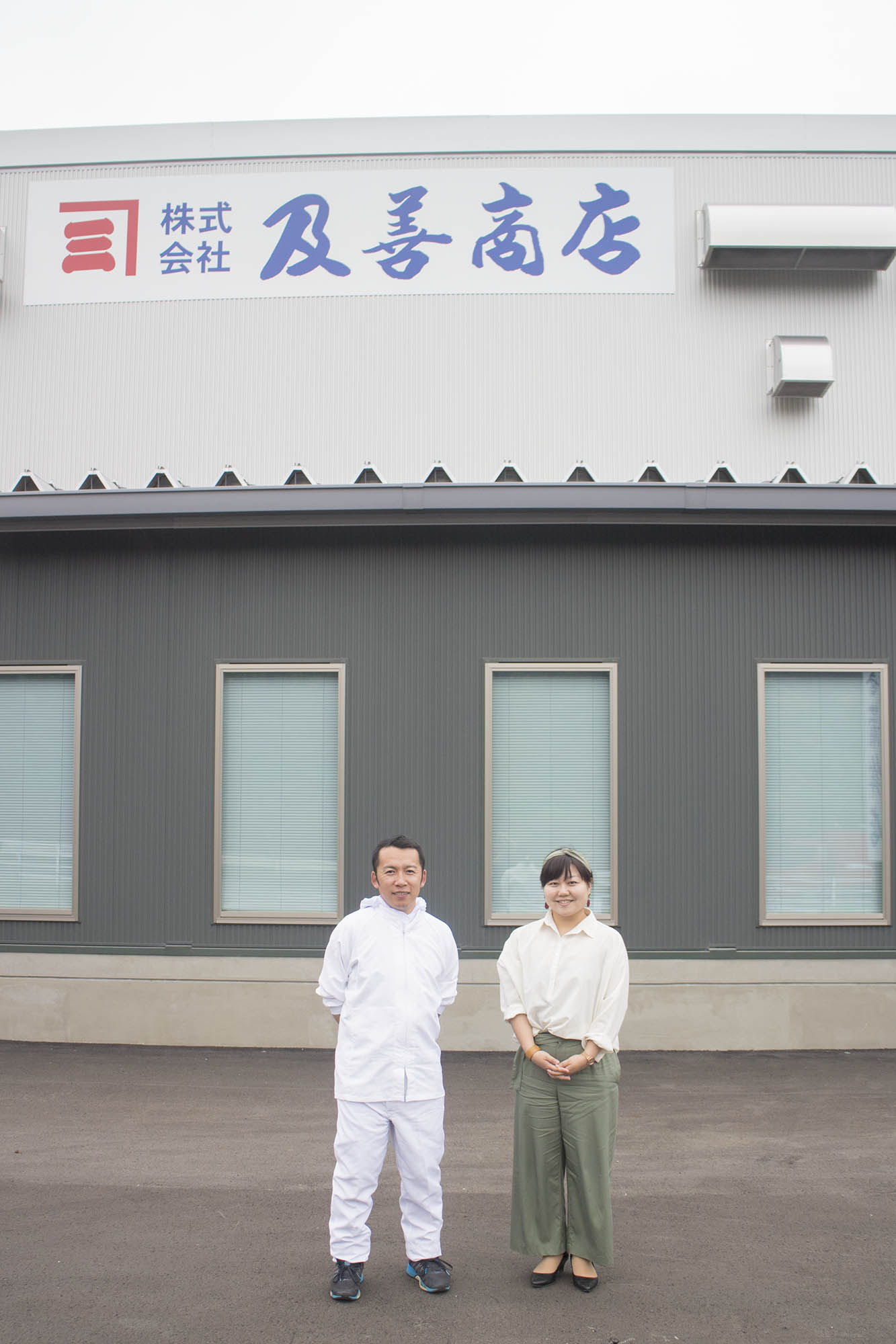 「株式会社 及善商店」と書かれた工場の建物の前で白い作業着を着た及川さんとリポーターの女性が並んで立っている写真