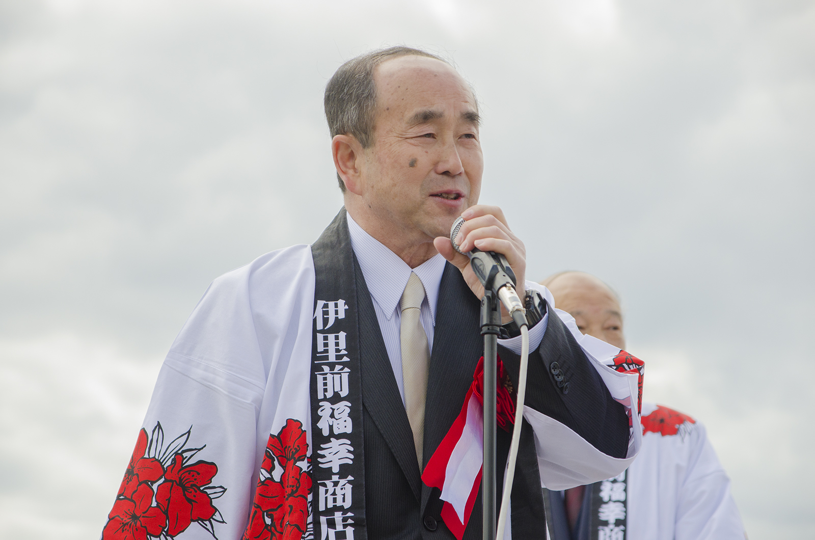 白地に赤い花の模様が入った法被を着た高橋武一さんがスタンドマイクを握って笑顔で話をしている写真