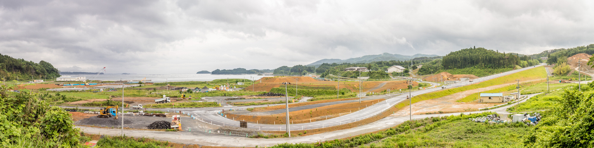 曇り空が広がる広大な海沿いの土地の定点カメラによる風景で左奥には、湾のような入り江が広がっており、小さな島や岬が遠くに見え海辺のすぐそばには仮設の建物やクレーン、重機が並び、海に向かって建設が進んでいる様子を写した写真