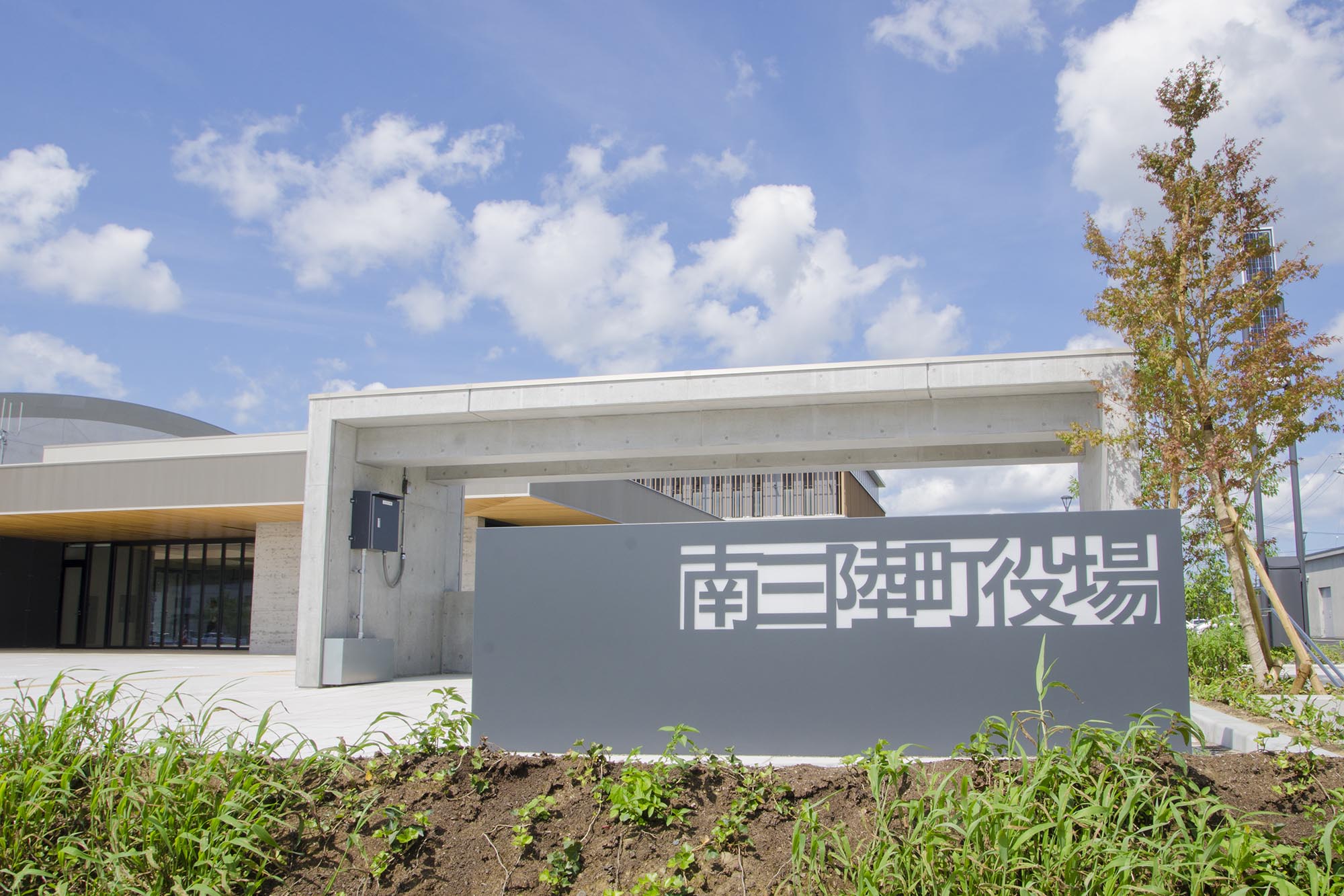 青空の下、コンクリート製の近代的な庁舎の前に「南三陸町役場」と大きく書かれた灰色の看板が設置されている写真