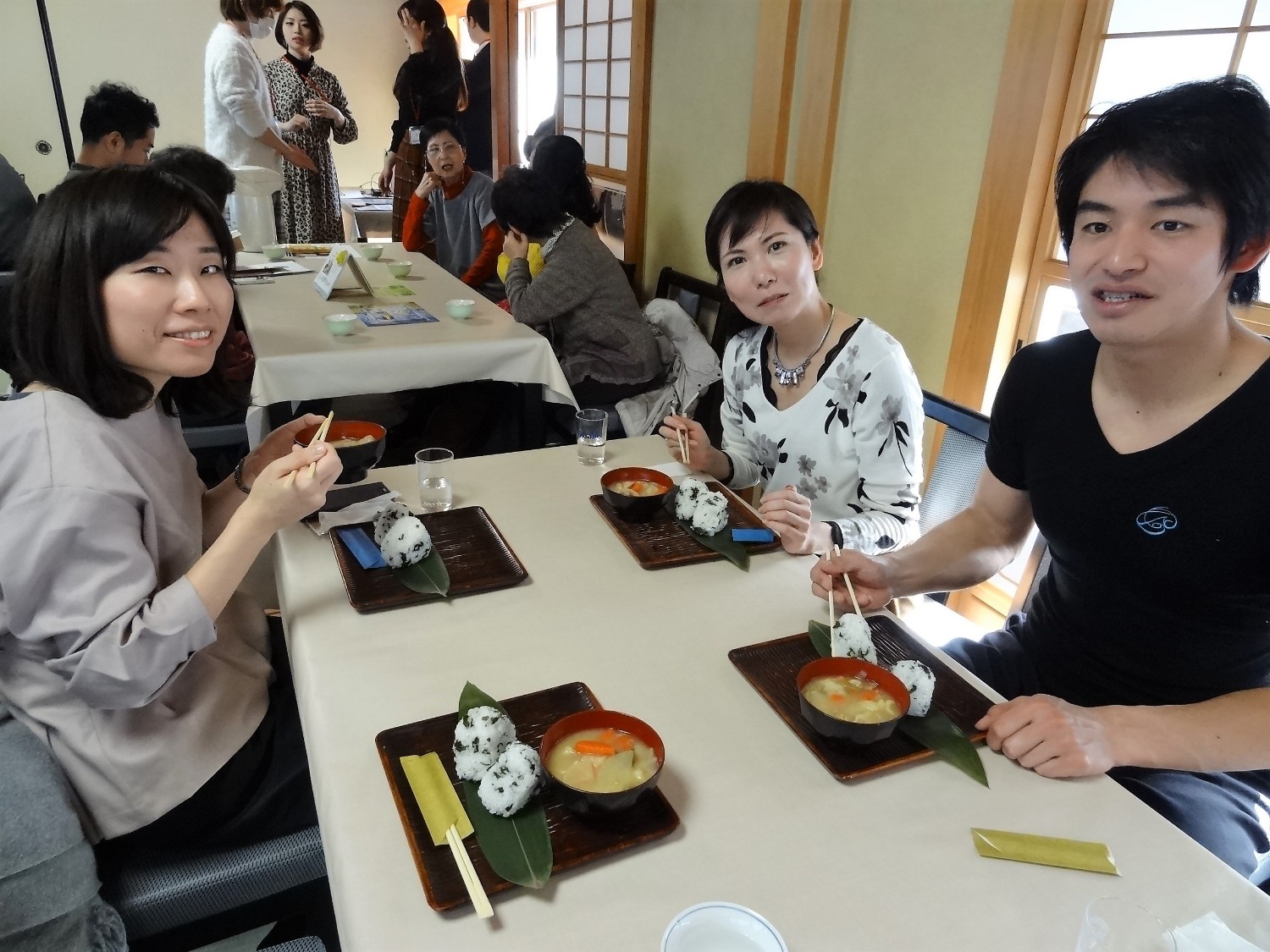 数人の人々が畳敷きの和室に座り、低い座卓を囲んで食事をしている写真