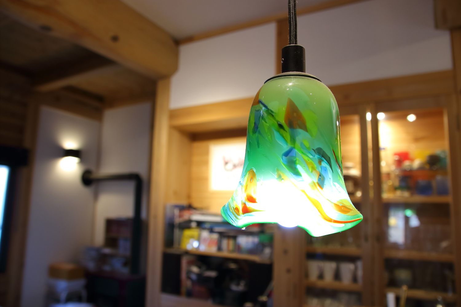 食器棚と木の壁が見えている店内に、天井に吊り下がった緑を基調に青やオレンジなどの模様が入ったガラス製のペンダントライトをアップで撮影した写真