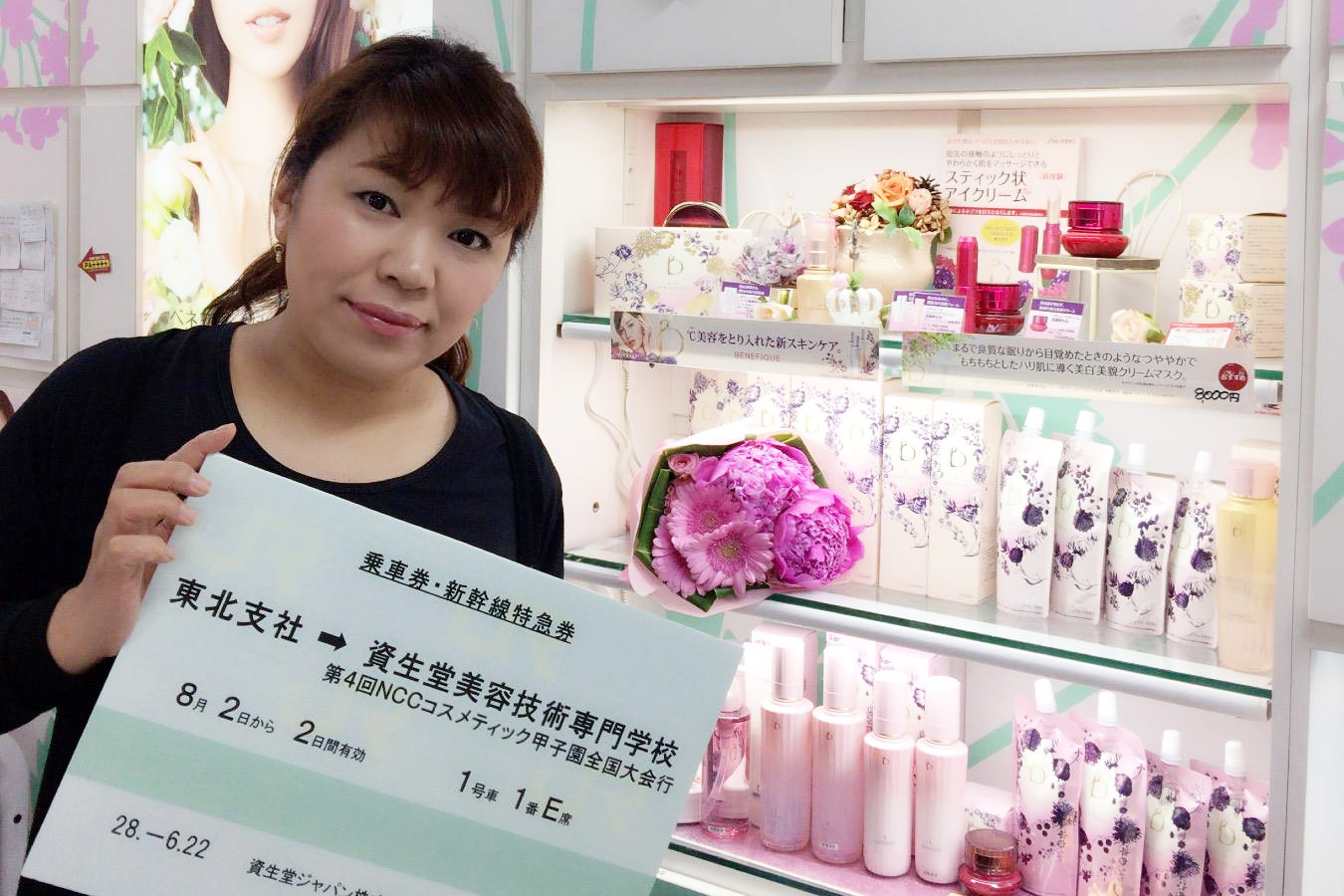 ピンクや白の化粧品が並んだピンクの花束の造花が飾られた化粧品棚の前で、黒い服を着た女性が「乗車券・新幹線特急券 東北支社から資生堂美容技術専門学校」と書かれた大きなパネルを両手で掲げている写真