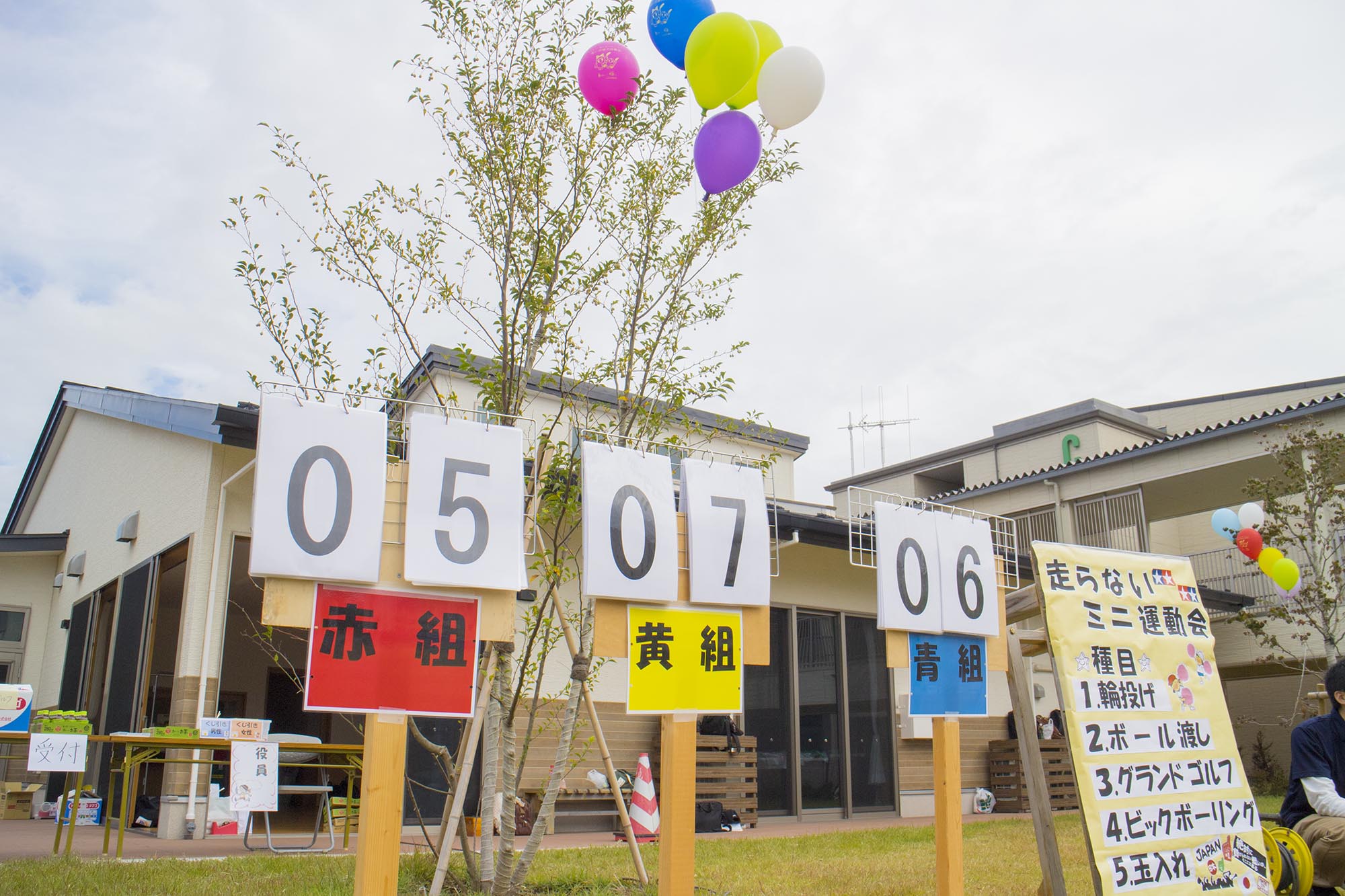 運動会の会場で赤組・黄組・青組の3チームの得点がボードに掲示され、背景には建物と風船の飾られた木があり、競技種目が書かれたの看板が置かれた様子を写した写真