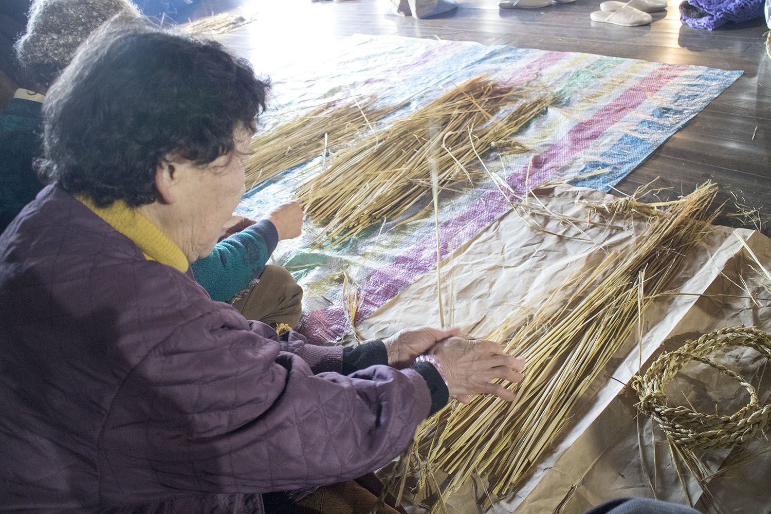 参加者が床に広げたシートの上で稲わらを束ねながら、丁寧に編み込んでわら細工を制作している様子を横から捉えた写真