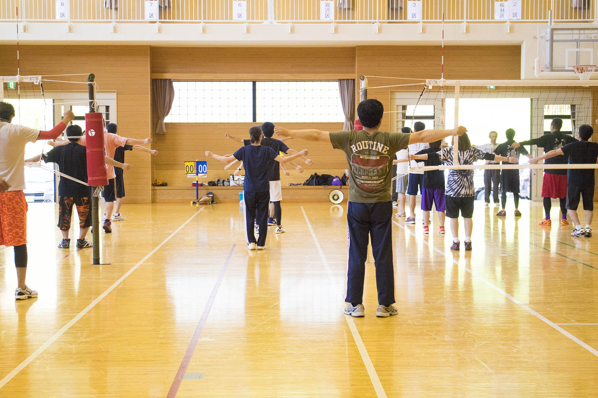体育館のバレーボールコートで、参加者たちが整列し、両手を横に広げて準備運動をしている様子を後ろから捉えた写真