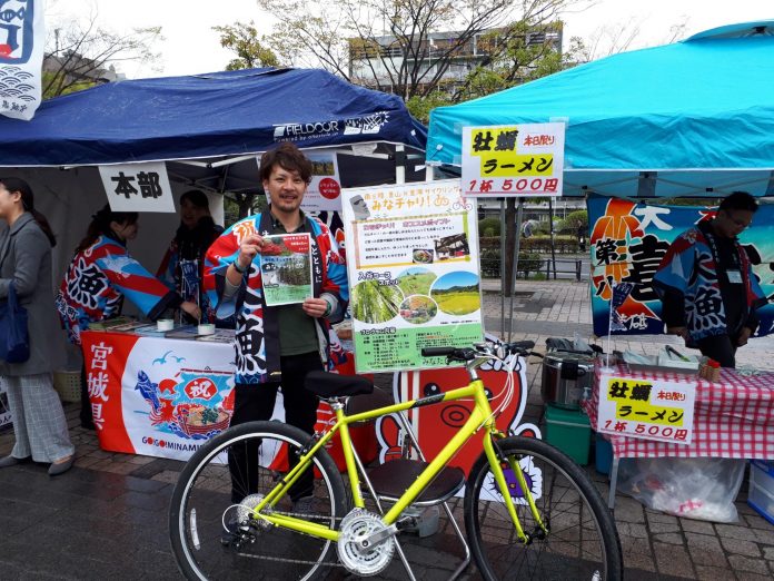 福興市会場の本部のあるテントの前にとめられた黄色の自転車の前で、チラシを持った男性が立ちPRを行っている写真