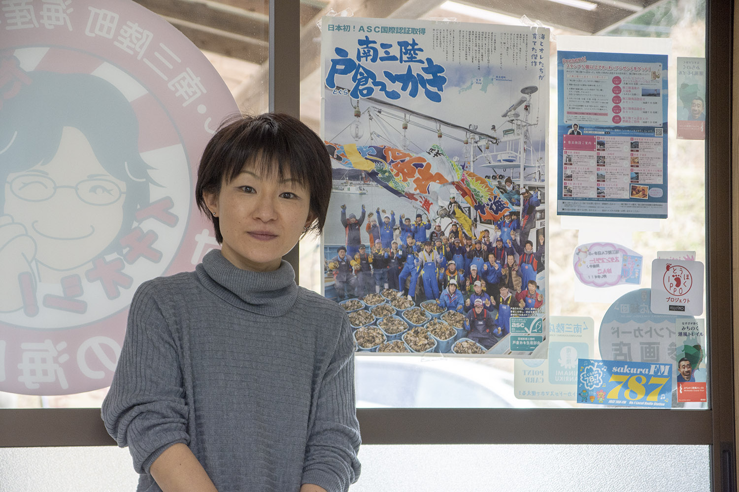 「南三陸戸倉っこかき」と書かれたポスターが貼られた窓の前に立ち、グレーの服を着た女性が真っすぐカメラを見て立っている写真