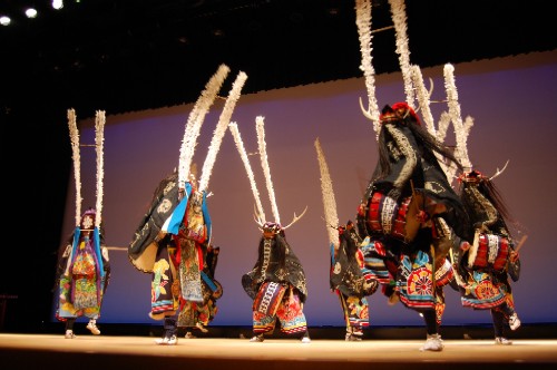 ステージ上で能面のような面と鹿の角、黒い衣装に派手な飾り付けを身につけ、背中に白い棒状の長い飾りを背負った複数の演者たちが、舞台上で力強く舞い踊っている様子の写真