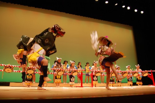 ステージで、獅子舞と鮮やかな衣装の演者が対峙して舞い、その後ろでは多くの子どもたちが太鼓を叩いている様子の写真