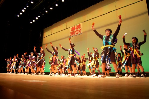 ステージ上で、お揃いの鮮やかな衣装を身につけた大勢子どもたちが、手に鳴子を持って両腕を力強く突き上げ、躍動感あふれる踊りを披露している発表会の様子の写真