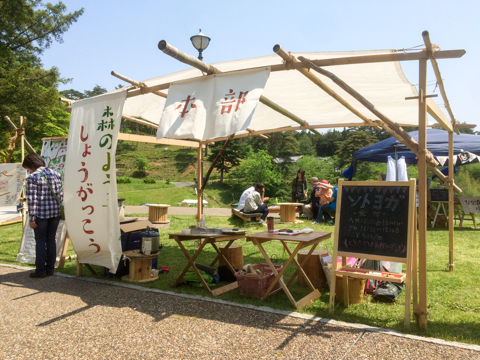 竹と布で作られた簡易テントに「本部」と書かれた布の看板、「森のようちえんしょうがっこう」と書かれた看板があり、後ろにはベンチやテントがある様子の写真
