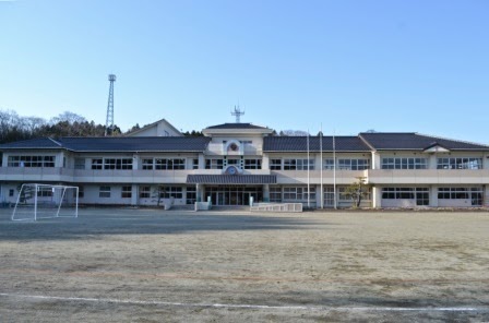 広い校庭の向こうに、時計台のある白い二階建ての校舎が左右対称に建っており、校庭にはサッカーゴールや旗ポールが見える晴天のもと撮影された小学校の写真