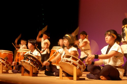 座った姿勢で鉢巻を締めた数人の子どもたちと、その後ろに立つ子どもたちが、ステージ上でそれぞれ和太鼓を力強く演奏している発表会の様子の写真