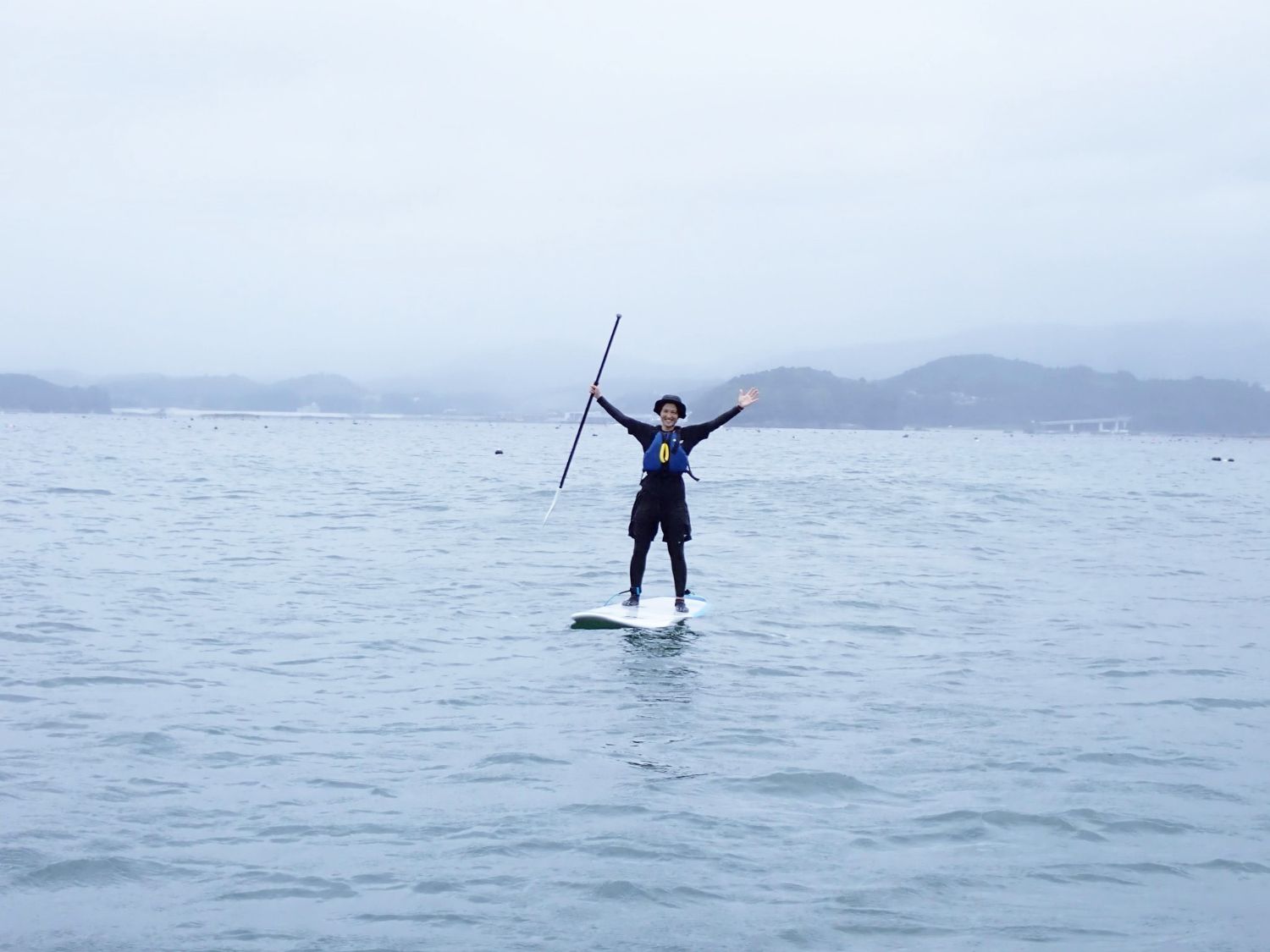 曇り空の海の上で、女性隊員がライフジャケットと帽子を身につけてスタンドアップパドルボードに乗り、両手を広げてポーズをとっている写真