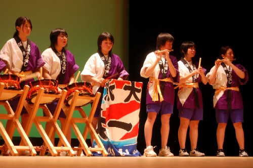 ステージ上で、法被を着用した複数の子どもたちが、太鼓や横笛演奏している発表会の様子の写真
