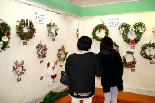 屋内壁面に、ハート型、動物やキャラクターをモチーフにしたクリスマスリースなど、様々なデザインのものが展示され、来場者がそれらを鑑賞している写真