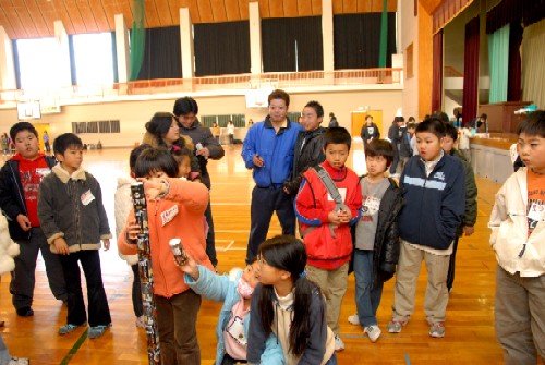 体育館で、子どもたちがゲームのようなものを囲んで集まり、大人たちが見守る中で楽しんでいるふれあいまつりの様子の写真