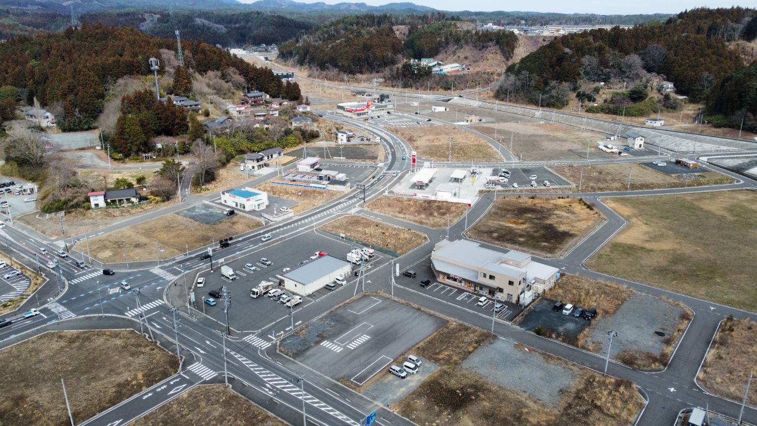 交差点を中心にコンビニや小規模な建物が点在し、周囲に山と住宅地が広がる志津川の郊外地区の昼間の風景を高台から定点カメラで撮影した写真