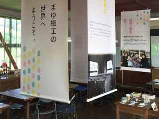 「まゆ細工の世界へようこそ」と書かれた垂れ幕が下がる教室のような室内で、繭を使った作品が展示されている写真