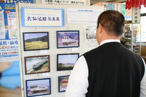 男性が、「気仙沼線 写真展」と題された掲示板に飾られた、列車と田園風景や海岸線などが写った鉄道写真を鑑賞している様子を背後から撮影した写真