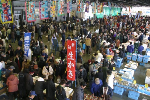 市場で、頭上や通路にカラフルな旗やのぼりが飾られ、大勢の客が集まり、青いコンテナに積まれた魚介類や商品を熱心に見て回っている、賑やかなおすばで祭りの写真