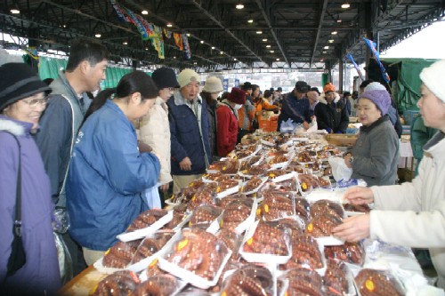 屋内の市場で、大勢の客が、手前のテーブルに山積みされた大量のパック詰めのタコを興味深そうに見ながら店員と会話しているおすばで祭りの写真