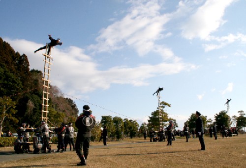 青空の下、広々とした屋外の広場で、はしごのような高い竹組の上で演技をする人々と、それを支え見守る関係者の写真