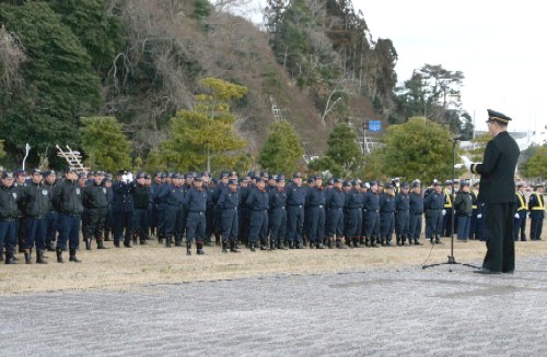 整列した多数の消防団員や関係者を前に、制服姿の幹部と思われる人物がマイクを使って訓示を行っている出初式の様子の写真
