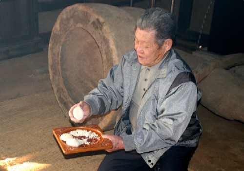 座っている高齢の男性が、丸い餅を片手に持ち、もう一方の手にも盆に乗せられた丸い餅を持って米作況を占っている様子の写真