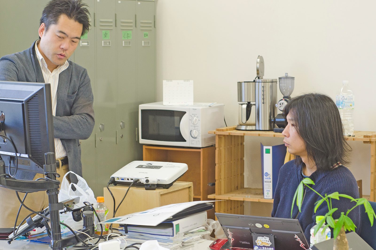 机の上には資料や飲み物が置かれているオフィスの一角で、パソコンの前で2人の男性が真剣な表情で会話している写真