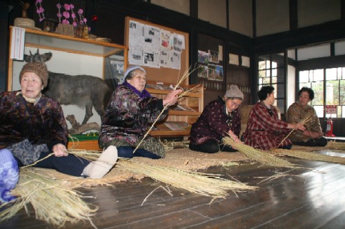 日本の伝統的な家屋の屋内で、数人の高齢の女性たちが、藁の束を使い、座って伝統工芸品のようなものを作っている様子の写真