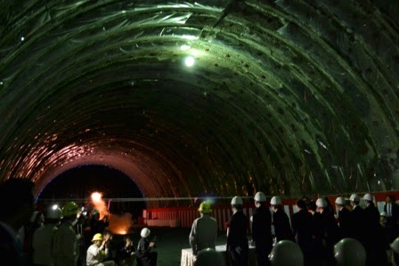 ヘルメットを着用した人々が一か所だけ灯りが灯された暗いトンネル内に集まり、カメラを構えていた左側に座っている人たちが後ろを振り返り、花火の様な炎が少し離れた地面から奥の壁に向かって発破している貫通式の様子の写真