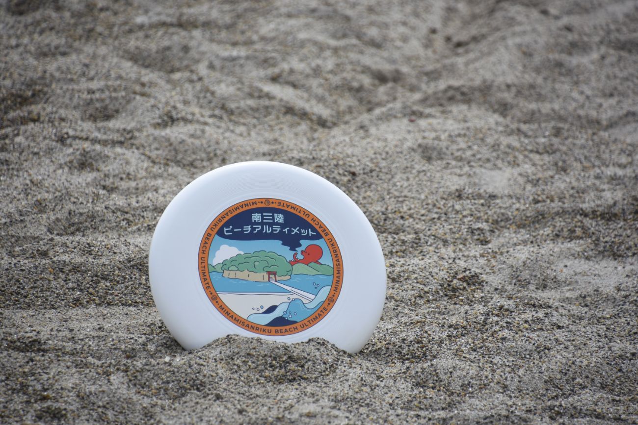 砂浜の上に立てられた白いフライングディスクの中央に、タコと島、波のイラストとともに「南三陸ビーチアルティメット」と書かれたロゴが印刷されているビーチアルティメット大会の記念ディスクの写真。