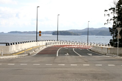 新たに舗装された道路が、湾の穏やかな水面を背景にカーブを描きながら海へ向かって下っているはまゆり大橋と遠くに見える椿島の写真
