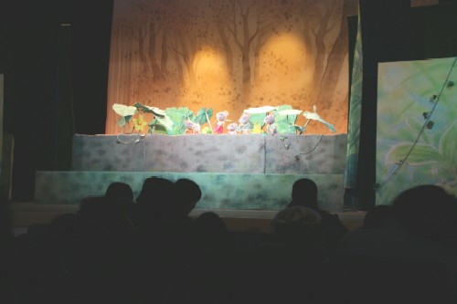 ステージ上で人形劇が行われているのを客席で多くの人が鑑賞している様子を背後から撮影した写真