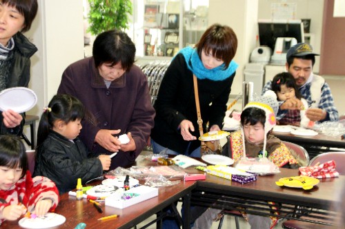 屋内で行われているワークショップで、数人の大人と子どもたちがテーブルを囲み、白い皿や色紙、接着剤などを使って、人形作りを行っている様子の写真