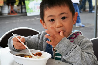 灰色のパーカーを着た小さな男の子がテーブルに座ってさんさんタコカレーをスプーンで食べながら、指についたルーをなめて満足げな表情をしている様子の写真