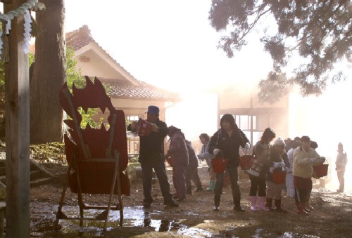 神社の敷地内で、多くの人々が赤いバケツを持って、大きな炎に見立てた的を狙って水をかけている訓練の様子の写真