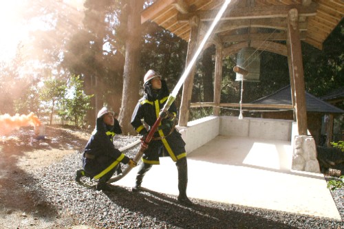 屋外で、消防服を着用した二人の関係者が、木造の鐘楼を背景に、ホースから勢いよく水を放水する訓練を行っている写真