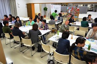 会議室のような明るい室内で、長机を囲んでグループごとに人々が活発に話し合っているグループディスカッションが行われている様子の写真