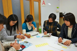 五人の女性が一つのテーブルを囲み、付箋やメモを使って真剣に意見をまとめたりノートをとったりしている写真