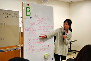 Bと表示されたホワイトボードの前に立つ女性が、マイクを持って赤字で書かれた意見を指差しながら発表している写真