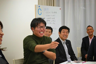 満面の笑みを浮かべた男性が指をさして話しながら他の参加者たちも笑顔で耳を傾けている、和やかな雰囲気の討議の様子の写真