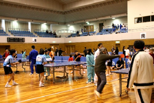 体育館で、多くの卓球台が並べられ、幅広い年齢層の人々が卓球の試合や練習に熱中している様子の写真