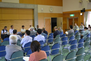 議室で多くの参加者が着席し、前方の机に座る関係者の方々が会議を進行している様子の写真