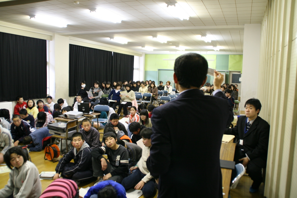 室内で、スーツ姿の気仙沼税務署の職員講師が、大勢の生徒たちに向かって話をしている様子を背後から撮影した写真