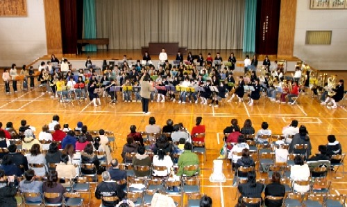 体育館の床に設けられたステージで、指揮者を中心にして、大勢の生徒たちが、管楽器を演奏する吹奏楽団としてパフォーマンスを披露しており、それを保護者などの大勢の観客が椅子に座って鑑賞している演奏会の写真